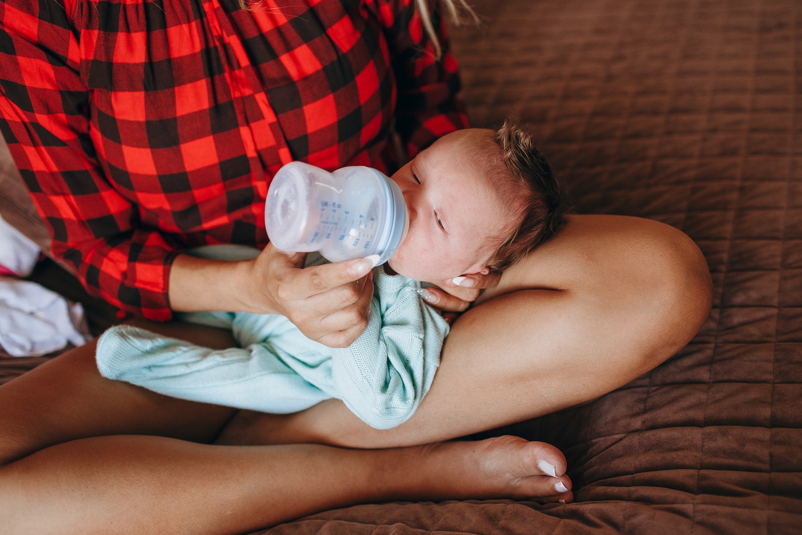 Crop anonymous mother feeding adorable newborn baby with bottle while sitting on bed with crossed legs in light bedroom at home